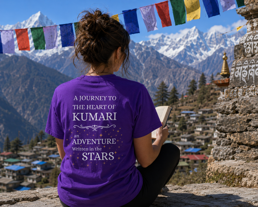 Person wearing a purple t-shirt with text, sitting on a stone wall with mountains and prayer flags in the background.